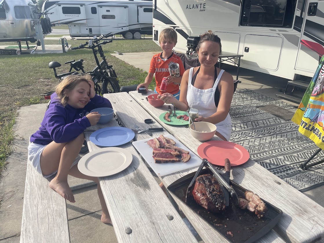 Two children and an adult preparing food at a picnic table outside an RV.