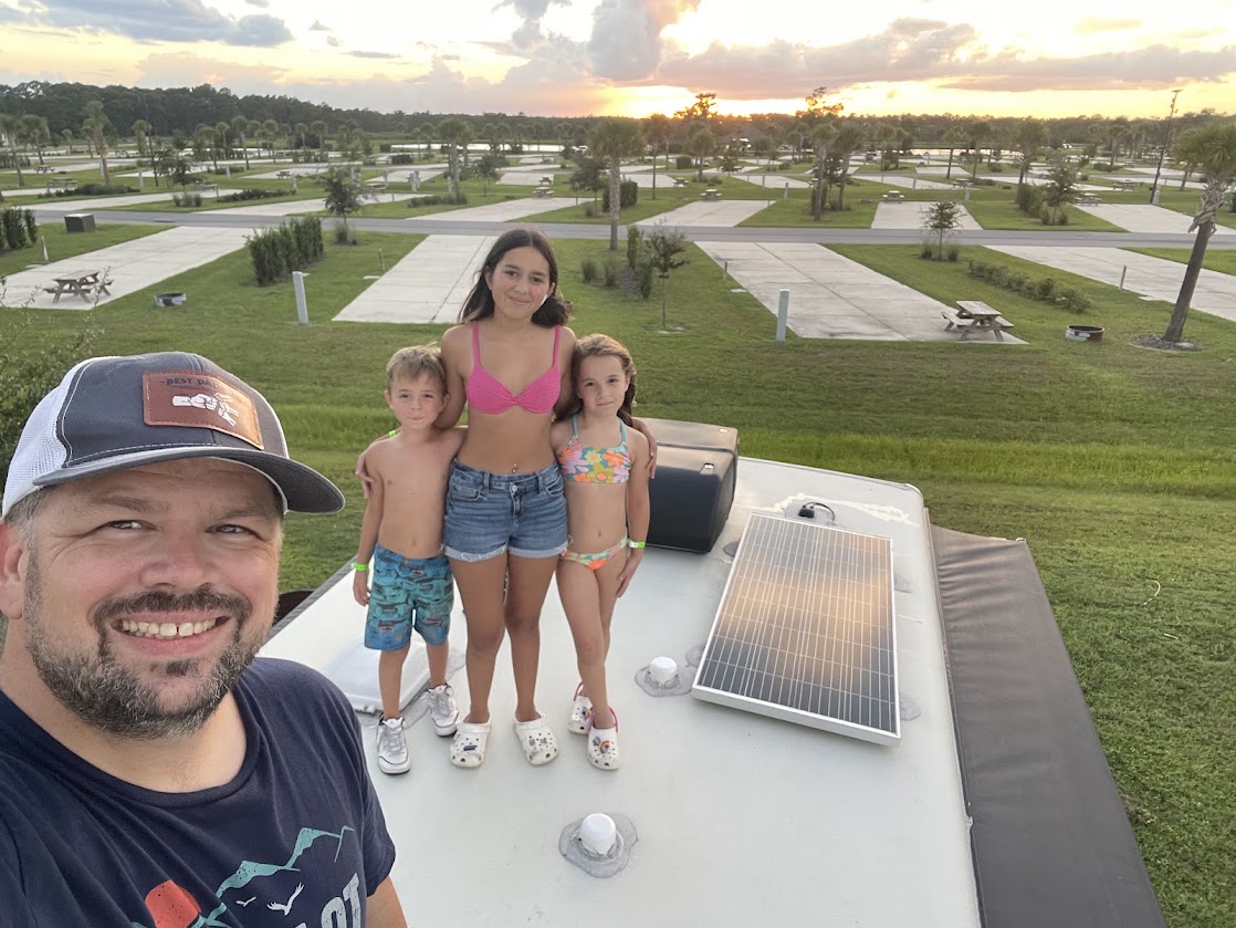 A man posing with two children on an RV roof, with a sunset in the background.