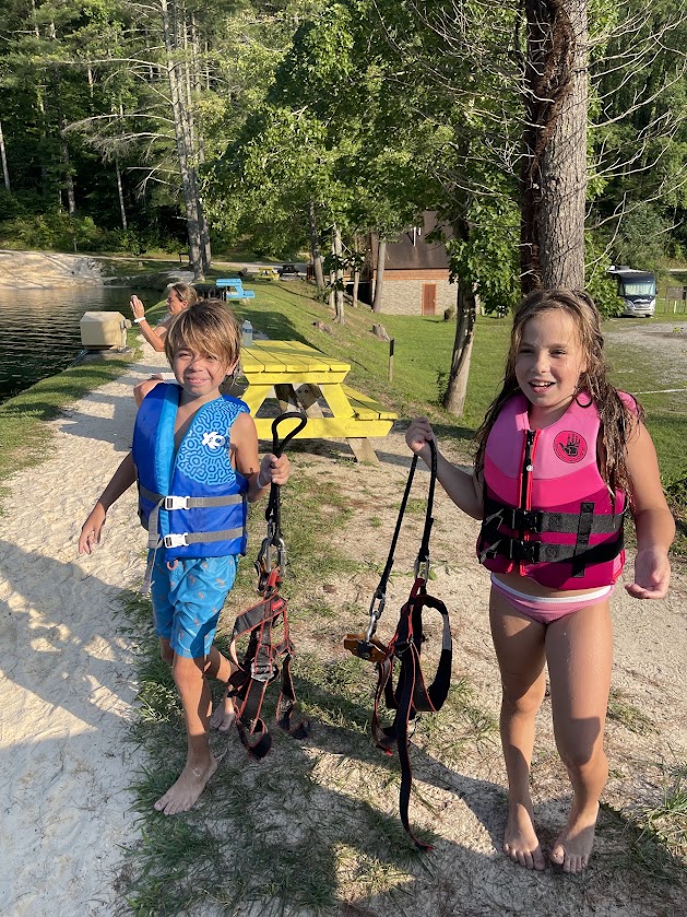 Two children wearing life jackets holding harnesses near a lake.
