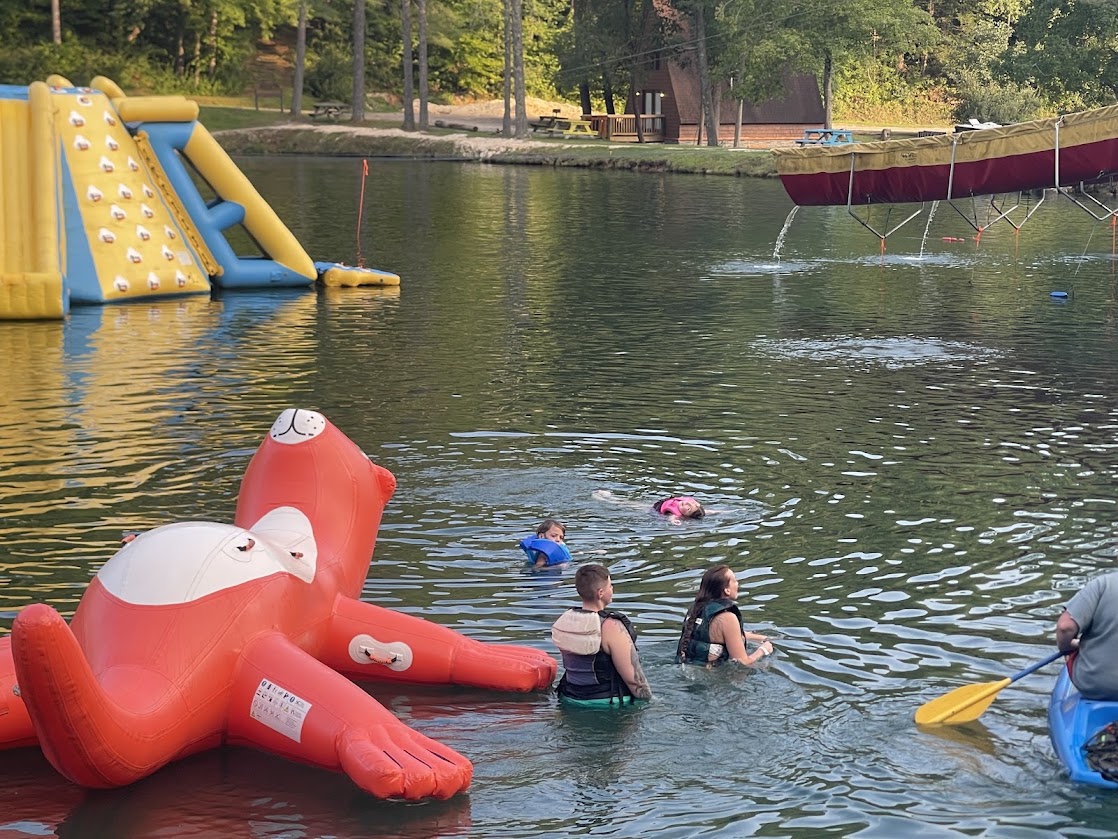People swimming in a lake with inflatable toys and kayaks visible.