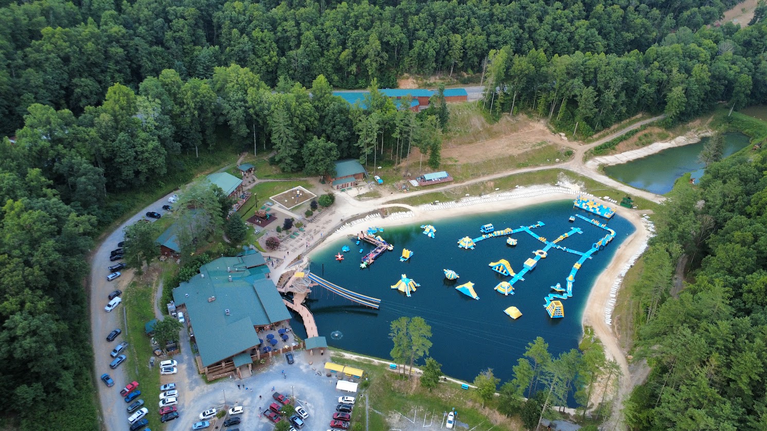 Aerial view of a lodge area with inflatable water attractions in a lake surrounded by trees.