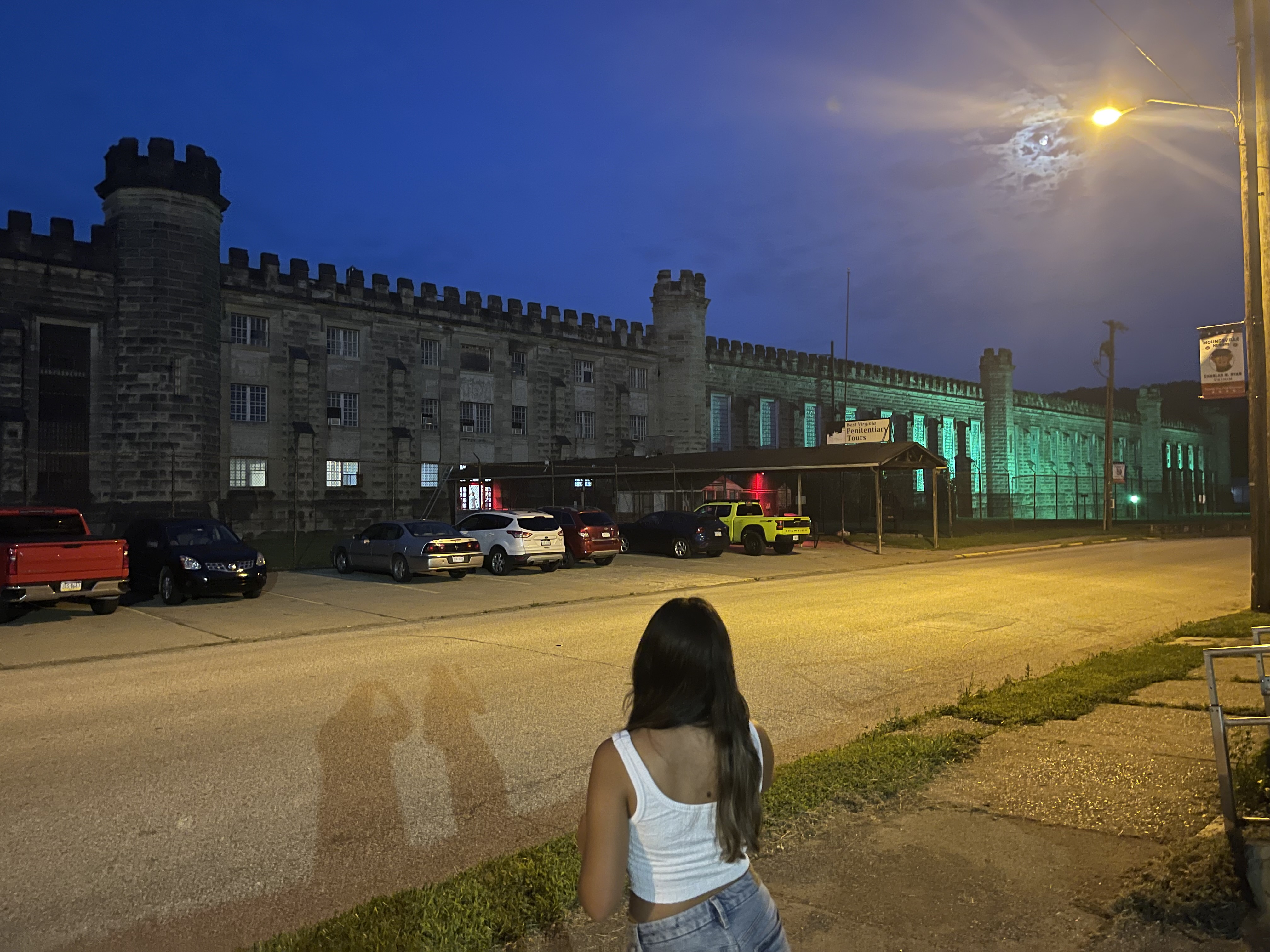 The West Virginia Penitentiary at night, illuminated by green lights with parked cars in front.