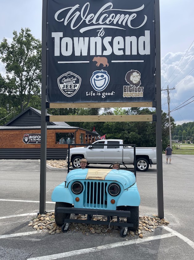 Two children sit in a blue Jeep under a 'Welcome to Townsend' sign.