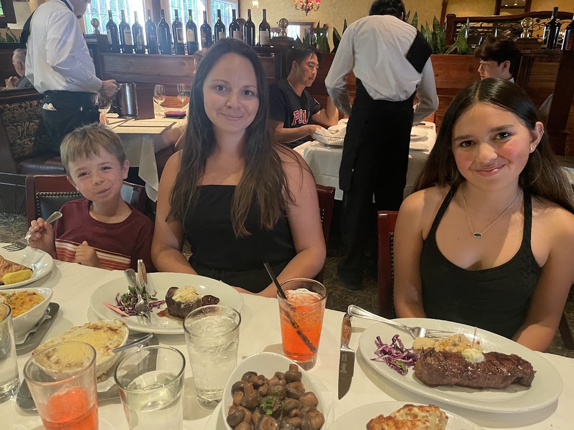 Three diners at a restaurant table with dishes including steak and sides.