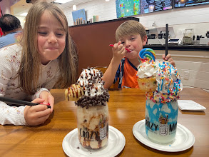 Two children sharing two elaborate milkshakes at a table.