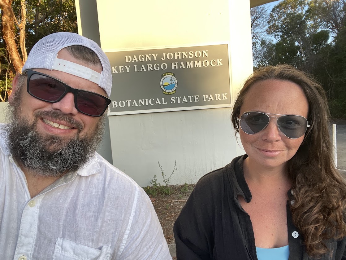 Two adults smiling in front of the Dagny Johnson Key Largo Hammock Botanical State Park sign.