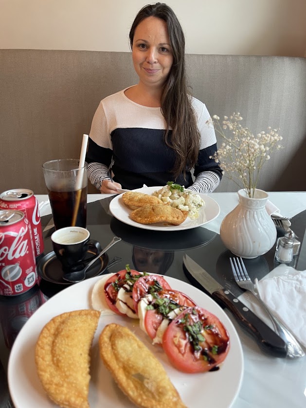 A woman sitting at a table with empanadas and a tomato salad in front of her.