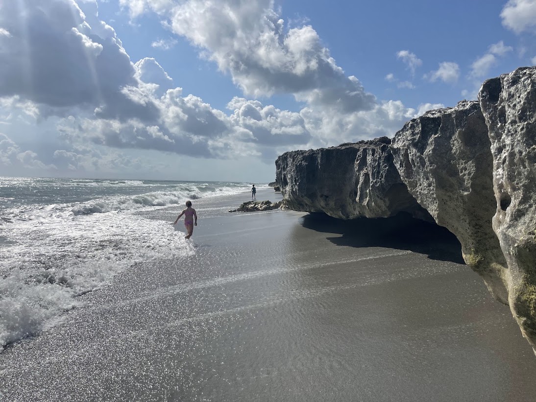 A child wading in the shallow surf near a large limestone rock on a sunny beach.