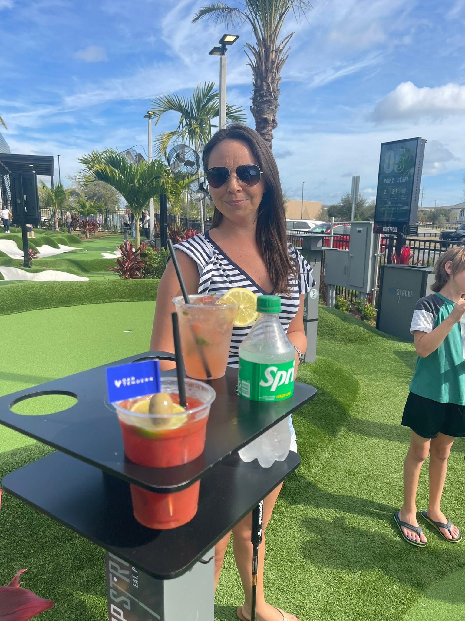 A woman stands beside a drink holder with cocktails and a soda at Pop Stroke mini golf.