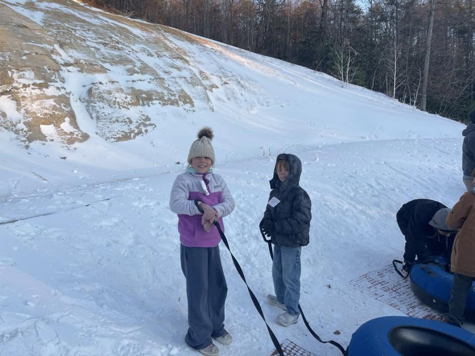 Two children stand on snowy ground near a snow tubing hill, smiling.