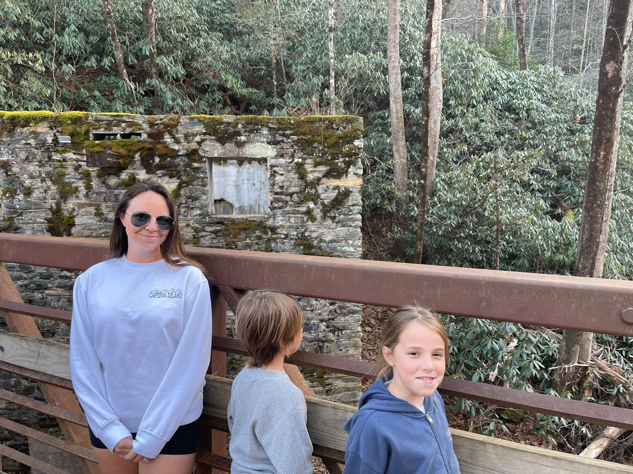 A woman and two children stand on a bridge by a stone ruin, surrounded by trees.