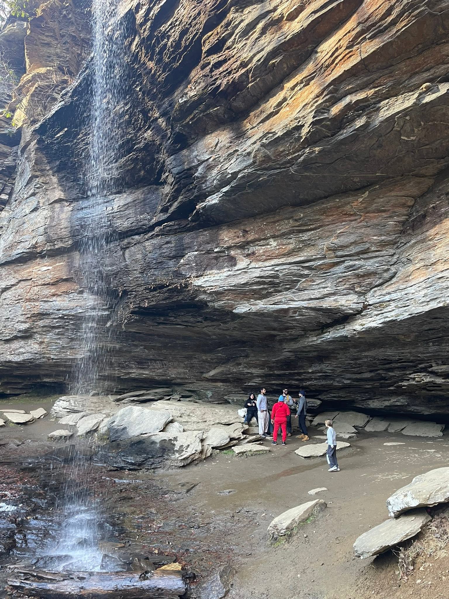 Group of people standing under Moore Cove Falls, with water cascading down rocky cliffs.