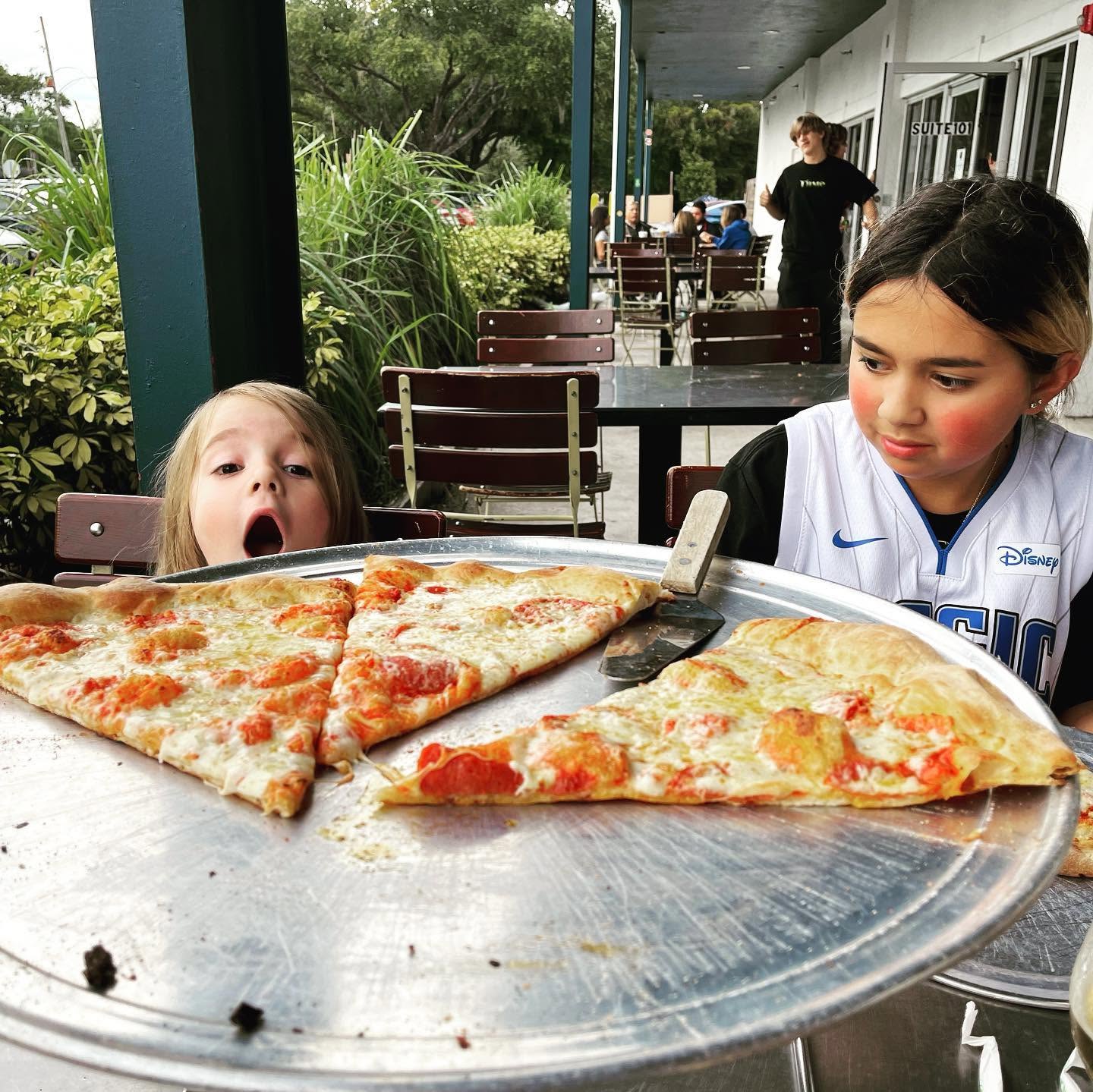 Two children at an outdoor table, excitedly watching large pizza slices.
