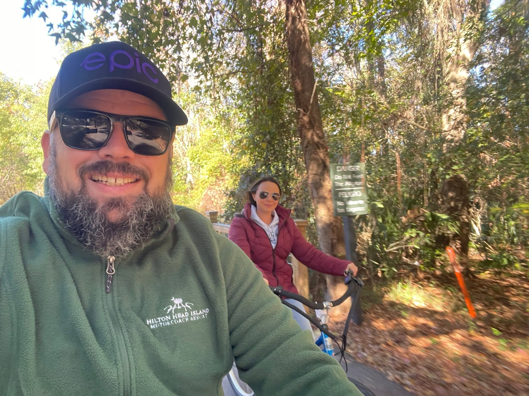 Two people biking on a wooded path near Hilton Head, with a warning sign visible.