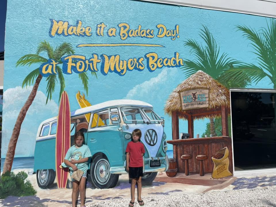 Two children standing in front of a colorful mural featuring a vintage van and tropical scenery.