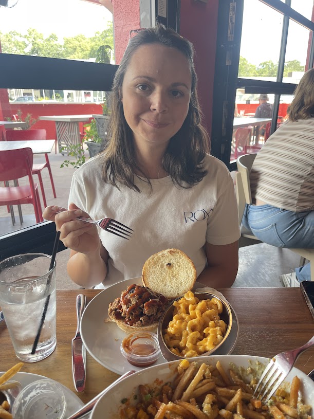 A woman holding a fork, posing with a sandwich and side dishes at Pigzza.